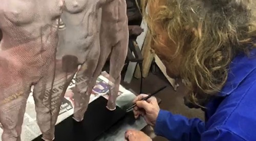 An artist is seen signing the black base of a rose-gold coloured wire sculpture depicting three girl figures